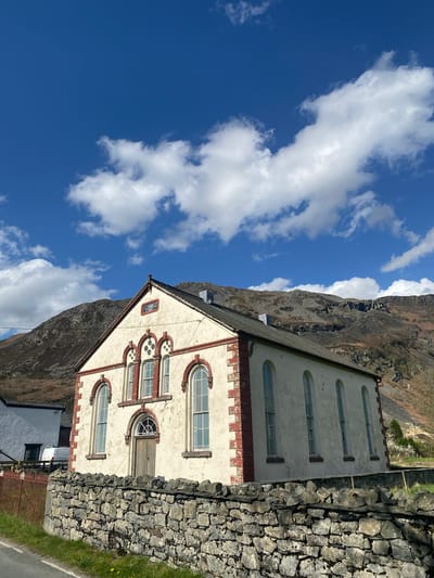 The image shows a building in Oswestry, United Kingdom, next to a rock wall and a mountain. The building is cream-colored with red brick trim and has multiple windows. The sky is blue with white clouds.