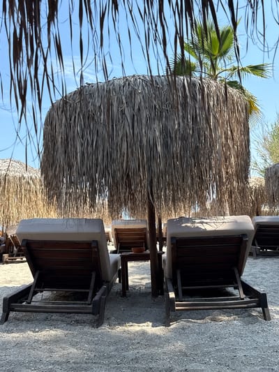 Two empty wooden lounge chairs with beige cushions are positioned on a sandy beach in Pallini Municipal Unit, Greece, facing away from the camera. Both chairs are placed under a large thatched umbrella made of dry palm leaves, casting shadows on the ground. A small wooden table sits between the two chairs, with a brown bowl visible on its surface. In the background, more thatched umbrellas and lounge chairs are visible, suggesting a resort or beach club setting. A palm tree can be seen behind the central umbrella, and the sky is clear and blue. The sand is a light grey color and appears to be composed of small pebbles and grains. The scene is peaceful and suggests a relaxing atmosphere, likely on a sunny day.