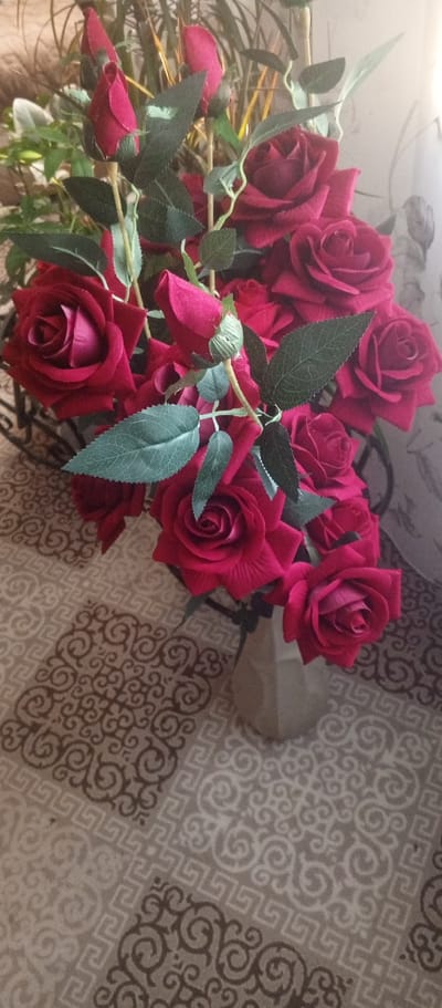 A bouquet of deep red roses and buds is displayed in a light-colored vase, placed on a patterned tablecloth. The roses are the primary subjects, with their velvety petals and green leaves filling most of the frame. Some roses are fully bloomed, showcasing their intricate spiral structure, while others are still in bud form, their rich color hinting at what's to come. The vase is simple and geometric, its muted tone allowing the vibrant roses to stand out. The tablecloth beneath has a repeating pattern of swirls and geometric shapes in earthy tones of beige and brown, creating a textured visual foundation for the floral arrangement. In the background, hints of a curtain and possibly other foliage can be seen, suggesting an indoor setting. The lighting appears to be natural and soft, casting gentle shadows and highlighting the depth and richness of the rose petals. There are no people or discernible activity taking place; the scene is a still life composition focused on the beauty of the flowers. The image evokes a sense of quiet elegance and natural beauty. There is no visible text within the image.