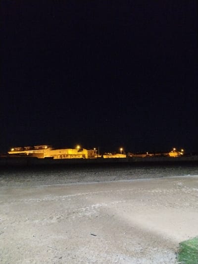 It's nighttime, and a lit building sits on the horizon. The ground in the foreground is sandy with a hint of grass at the bottom right. The sky is dark and clear, with two small dots of light towards the top. The building has multiple sections and appears to be a residential area. Streetlights are visible along the road.