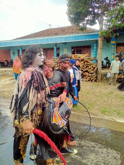 The scene depicts an outdoor gathering on a wet paved surface in Kedawung, Indonesia, against a background of a light blue building with a brown tiled roof and a stack of wood. Three central figures are prominent.
On the left, a person with pale white face paint, dark disheveled hair, and a tattered, multi-layered dark and light-colored costume is visible. This individual wears brass bells on the ankles and holds a thick red braided rope in the right hand. Their mouth is open, and they are looking towards the right.
Next to this figure, a second individual has a red, contorted face, an open mouth, and appears to be crying or in an altered state. This person is wearing a dark, wet shirt with red lettering and is being supported by the third central figure.
The third central figure, positioned to the right of the second, has a dark complexion, a patterned headband, and red and dark markings on the face. This person wears a dark, wet long-sleeved shirt with red accents and holds a thin black whip-like object in the left hand, while supporting the second individual with the right arm.
Between the first and third figures, at waist height, is a black, stylized flat horse effigy with orange, red, and white patterns, consistent with a Kuda Lumping performance.
In the background, several bystanders are visible: a woman in an orange headscarf walks on the left, a woman in a blue headscarf holds a child in the center, and a man in a light green shirt carries a bag on the right. A large pile of cut logs is stacked against the building in the mid-ground. The ground in the foreground is wet.