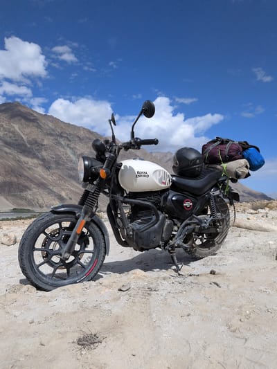 A Royal Enfield Hunter 350 motorcycle is parked on a rocky, sandy terrain. The motorcycle is white with "ROYAL ENFIELD" and "HUNTER" logos visible. It is equipped with saddlebags packed with camping gear, including a helmet. The background features arid, mountainous landscape with a clear blue sky dotted with white clouds. The location is identified as Sasoma, India.