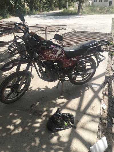 A stationary, maroon and silver motorcycle is parked on a concrete surface under dappled sunlight. The motorcycle has a black seat, a black luggage rack on the rear, and a black front fender. A black plastic trash bag, partially filled with items, is on the ground in front of the motorcycle. In the background, a metal gate, some construction materials, and a building are visible. The scene is located in Danghara, Tajikistan.