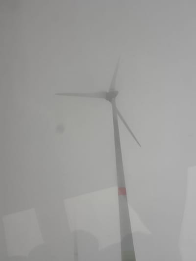 A single wind turbine is depicted partially obscured by dense fog in Schöpstal, Germany. The turbine's dark grey central tower rises vertically, exhibiting a prominent rectangular red band on its lower segment. Three blades are visible, extending from the nacelle into the heavy, monochromatic grey fog which uniformly dominates the sky and background, severely limiting visibility. The lower portion of the image contains multiple light-colored, rectangular reflections, consistent with internal objects or light sources, superimposed over darker, amorphous shapes, indicating the image was captured through a reflective surface, likely a window. No discernible individuals or specific actions are present within the scene.