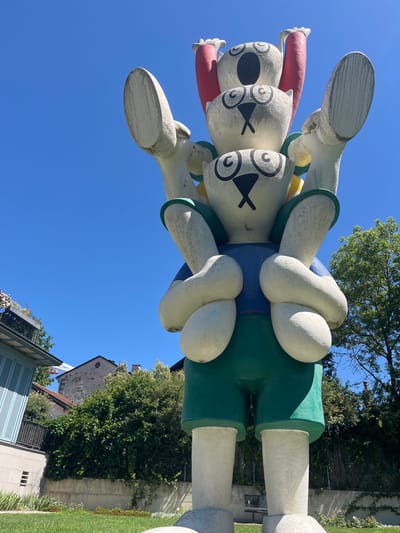 This image captures a vibrant, larger-than-life sculpture in Cologny, Switzerland. The sculpture appears to be of stylized figures, possibly cartoon characters, with oversized, rounded limbs and distinct facial features. These figures are painted in bright colors, including white, red, yellow, blue, and green, suggesting a playful and whimsical subject matter.
The sculpture is viewed from a low angle, looking up towards a clear, bright blue sky, indicating it's a sunny day, likely in the late morning or afternoon. Lush green trees and foliage frame the top and right sides of the image, with a glimpse of buildings in the background. The architecture of the buildings, with their stone facades and pitched roofs, adds to the European setting. The overall atmosphere conveyed is cheerful and engaging, possibly part of an outdoor art installation or a public park. There is no visible text in the image.
