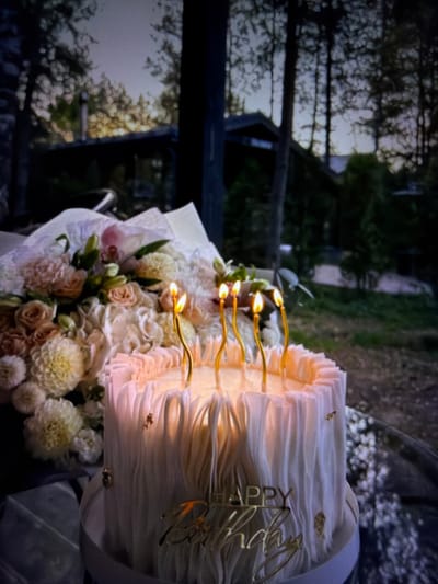 The image shows a birthday cake with five lit candles and a "Happy Birthday" cake topper. Next to the cake is a bouquet of flowers. The cake and flowers are placed on a reflective surface. In the background, there is a house nestled amongst trees, suggesting an outdoor setting in a wooded area. The lighting indicates it is either dusk or dawn. The location is identified as Beșalma, Moldova. There are no people visible in the image.