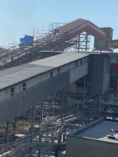 The image shows a tall industrial structure, likely under construction or undergoing maintenance, in what appears to be a facility near Poland. The structure is characterized by a complex network of scaffolding and metal beams, suggesting ongoing work. A prominent feature is a long, sloping conveyor belt system, partially covered with a corrugated metal roof. The conveyor belt is connected to a towering section of the facility, which has a red, angular roof or housing.

Against the clear blue sky, a bright blue tarp or umbrella is visible, snagged on the scaffolding. This detail might indicate a temporary covering for equipment or a worker's protection. The overall scene is one of industrial activity, with no people clearly visible. The lighting suggests it is daytime, likely morning or afternoon, with no strong shadows indicating a specific time. The presence of scaffolding and the incomplete nature of some parts of the structure point to a construction site. The industrial setting, combined with the Polish location context, suggests a manufacturing plant, power station, or similar large-scale industrial complex. There is no visible text in the image.