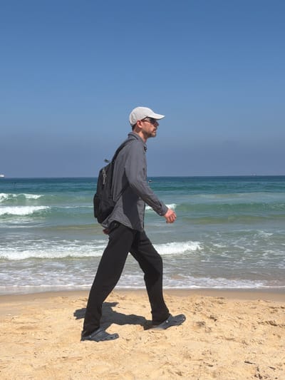 A man is walking on a sandy beach in Ashkelon, Israel. He is wearing a grey long-sleeved shirt, black pants, a white baseball cap, and sunglasses. He has a black backpack on. The sea is to his left, with waves breaking on the shore. The sky is clear and blue. In the distance, a ship is visible on the horizon. The sand is light brown and appears dry. The man is facing to the right, in profile.