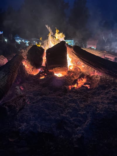 The image depicts a bonfire at night in Rapla, Estonia. Several large logs are burning, with flames and embers glowing brightly. Smoke rises from the fire into the dark sky. In the background, indistinct figures of people can be seen gathered around the bonfire, with some seated and others standing. Tents or shelters are also visible in the distance, suggesting an outdoor event or gathering. The ground in the foreground is covered in ash and debris from the fire. The overall atmosphere is warm and inviting, with the fire providing a focal point of light and activity in the dark.
