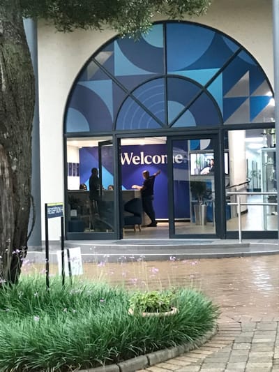 An external view of a building entrance in Midrand, South Africa, is depicted. The primary feature is a large, arch-shaped glass facade above the main entryway, segmented by dark frames and featuring geometric patterns in various shades of blue, white, and light grey. Below this, two glass doors lead into the building, with the left door slightly ajar.
Inside the building, a reception area is visible. A prominent blue wall behind the reception desk displays a large white "Welcome" sign. One individual, a light-skinned male wearing a dark top and trousers, stands in front of the reception desk, with his right arm extended slightly. A second individual, seen in silhouette, stands further left behind the open door near the reception desk. Reflections on the glass reveal internal office elements, including a television screen displaying a person, a potted plant, and metallic railings.
To the left of the building entrance, part of a large tree trunk and some foliage are visible. In the immediate foreground, a manicured garden bed contains long, green, grass-like plants and scattered delicate purple flowers on thin stalks, along with a small potted plant. A black signpost within this garden bed points right with "RECEPTION ->" printed in white. The area directly in front of the building is a brick-patterned paved surface, appearing wet, with three steps leading up to the entrance doors.