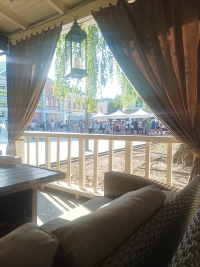 This image depicts an outdoor scene viewed from a covered seating area, likely a cafe or restaurant patio. In the foreground, a wicker table and a plush, grey cushion on a wicker chair are visible, suggesting a comfortable resting spot. The scene is bathed in bright sunlight, casting distinct shadows on the tiled floor, indicating it is daytime.
Beyond the seating area, a street scene unfolds. A white railing separates the patio from a paved area where many people are gathered. They appear to be participating in an event, possibly a race or a public gathering, with some individuals in athletic wear. In the background, white tents are set up, suggesting a market, festival, or similar outdoor event. Buildings with traditional architecture line the street, and trees with lush green foliage add to the ambiance. A lamppost with an ornate lantern hangs from the covered ceiling, adding a decorative touch.
The overall mood is one of a lively summer day in a public space. The bright sunlight and the gathering of people suggest a positive and engaging atmosphere. Some text is visible on a banner in the background, but it is not clearly legible. The location is identified as Zhytomyr, Ukraine.