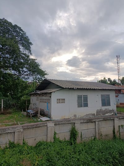 The image depicts a scene in Ban Patan, Thailand, featuring a modest white building with a corrugated metal roof, characteristic of rural or semi-urban dwellings. The building has two windows with louvred shutters, suggesting a tropical climate. Adjacent to the main structure is a smaller, open-sided annex with a similar roof and ventilation blocks, indicating it might serve as storage or a utility area. In front of the buildings, a concrete wall with some visible damage and vegetation growing along its top separates the property from a foreground of lush green grass.
Behind the buildings, a large, leafy tree dominates the left side of the frame, providing shade and a sense of natural surroundings. In the distance, a tall telecommunications tower stands against a cloudy sky, hinting at the presence of modern infrastructure. The sky itself is overcast with dramatic cloud formations, suggesting either the approach of rain or the aftermath of a storm. The lighting indicates it is likely late afternoon or early evening, with the sun attempting to break through the clouds.
There are no people visible in the image. Various objects are present, including what appears to be discarded materials and plumbing fixtures near the annex, suggesting ongoing construction, maintenance, or simply clutter. The overall impression is one of a lived-in, functional space in a rural or developing area, under a dynamic sky. No text is visible in the image.