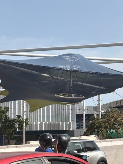 The image shows a wide shot of a modern outdoor structure with a dark gray fabric canopy. The canopy is supported by white poles and has a translucent blue section. A circular window with white spokes is visible on the canopy. In the foreground, there are parts of two cars, one red and one silver, and two people wearing motorcycle helmets. The person in the silver car is holding a phone. Behind the structure, there is a building with glass windows and horizontal white and blue stripes. A tree with small red berries is also visible. The sky is clear and blue, suggesting a daytime setting. The overall scene appears to be a street or public area in Palma, Spain, possibly during a sunny day. The presence of helmets and cars indicates people are likely traveling or taking a break.