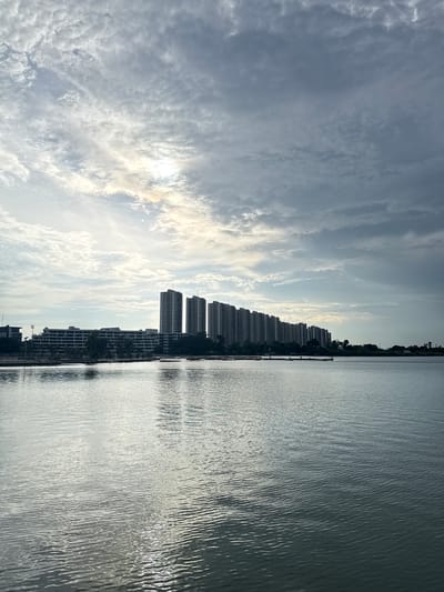 A wide-angle, eye-level shot captures a serene waterfront scene in Pak Kret City Municipality, Thailand, under a dynamic sky. The composition is bisected by the calm expanse of a body of water, reflecting the muted tones of the sky and the imposing structures that line the opposite shore.

Dominating the horizon is a series of modern, high-rise apartment buildings, stretching horizontally across the frame. Their rectangular forms and numerous windows are silhouetted against the clouds, creating a stark contrast with the natural elements. A lower, longer building with a more horizontal design is situated in front of some of the taller structures, featuring multiple floors with visible balconies or walkways. Trees and smaller buildings are interspersed between the residential blocks and along the waterfront, adding a touch of greenery and human habitation to the urban landscape.

The sky is a dramatic canvas of swirling clouds, with patches of sunlight breaking through, creating bright areas and subtle atmospheric effects. The sun itself is partially obscured, casting a gentle glow that illuminates the scene. The water in the foreground is slightly rippled, with sunlight creating shimmering patterns on its surface. The overall atmosphere is one of tranquility, with the urban environment harmonizing with the natural beauty of the sky and water. No individuals are visible in the frame, emphasizing the quietude of the moment.