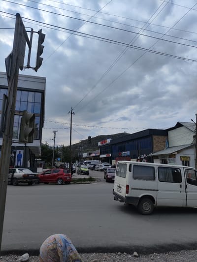 The image captures a street scene in Kerben, Kyrgyzstan, under an overcast sky. A traffic light stands prominently on the left side of the frame, with its signals facing away from the viewer. In the foreground, a white van is parked on the right side of the road. Behind the van, several other vehicles, including a red car and a silver car, are visible. A building with large windows and a modern facade is situated on the left, and further down the street, commercial establishments with signage are present. A person, partially visible and wearing a head covering, is in the lower foreground. The overall impression is a typical day in a town with moderate traffic and a mix of residential and commercial buildings.