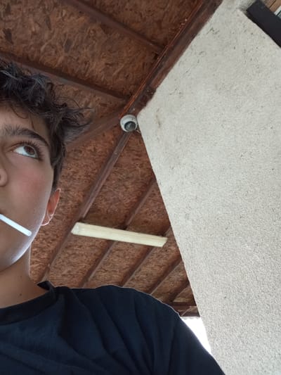A low-angle close-up shot depicts a person with dark hair and brown eyes, holding a white object, possibly a straw or lollipop stick, in their mouth. The person is wearing a dark-colored shirt. Above them is a ceiling constructed with brown beams and panels, likely made of wood or a similar material. A cylindrical security camera is mounted on the ceiling. Next to the ceiling is a textured, light-colored wall or pillar. The location cannot be determined from the image alone. The city of Gavrailovo, Bulgaria, is not evident from the visual information available.