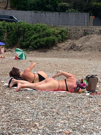 In Pozzarello, Italy, two individuals are sunbathing on a pebbled beach. The person in the foreground, lying on their stomach, has their legs extended and is wearing a black bikini bottom and a colorful headscarf. To their left, another person is lying on their stomach, facing away from the camera, wearing a black bikini. A dark-colored bag is placed near the person with the headscarf. Behind them, a green and white umbrella is partially visible, along with other beach accessories. The background features dense green foliage, a wooden fence, and a glimpse of a vehicle.