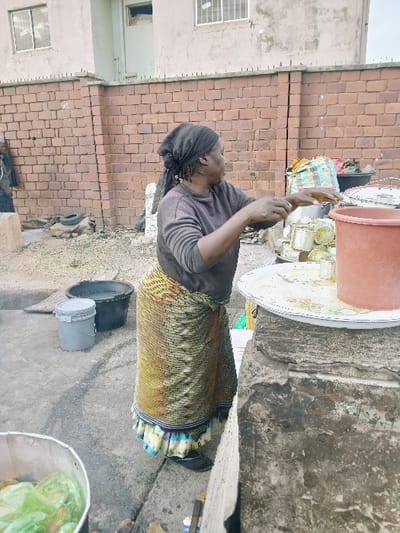 An older woman stands outdoors, engaged in food preparation. She wears a head covering, a long-sleeved shirt, and a patterned skirt. She is using her hands over a large tray filled with small, possibly wrapped, portions of food. Adjacent to her are containers of varying sizes and colors. The scene is set against a brick building.