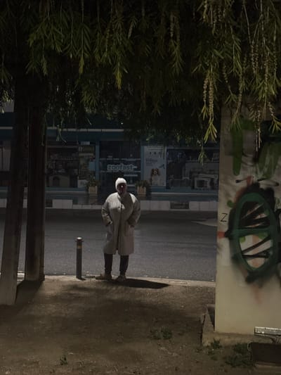 The image captures a nocturnal street scene in Egkomi Lefkosias, Cyprus. On the left, dense green foliage and bushes partially obscure the view. Above the foliage, a light-colored wall features graffiti, including a prominent green and black abstract, eye-like shape with internal lines, and a visible letter 'N'.

In the mid-ground, an adult person stands on what appears to be a dirt or gravel path, adjacent to a paved road. They are wearing a light-colored, long coat or jacket, dark pants, and a white or light-colored cap/hood. The person is facing slightly away from the camera, looking towards the background. A long shadow stretches from the person towards the right, indicating a light source behind them. A dark, short bollard is visible on the edge of the paved road near the person.

Across the dark asphalt road, in the background, are commercial buildings with storefronts. Artificial lighting from these establishments illuminates parts of the scene. Clearly visible text on one of the storefront signs reads "Zoofast", with "B.Vild" visible beneath it. Other text on signs is present but illegible due to darkness and resolution.

The time of day is night or late evening, characterized by the dark sky, artificial lighting, and pronounced shadows. The overall atmosphere is quiet and somewhat solitary.