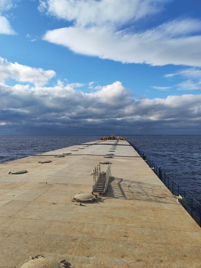The image shows a long, concrete pier extending into the water. The pier has a series of access hatches and a metal staircase leading down to the water. There's blue railing along the right side of the pier. At the end of the pier, there are some yellow objects, possibly buoys or equipment. The water is dark and choppy. The sky is filled with cumulus clouds, with patches of blue visible above. A layer of storm clouds looms behind the pier. It is assumed this is taken in Pavlokovo, Russia.
