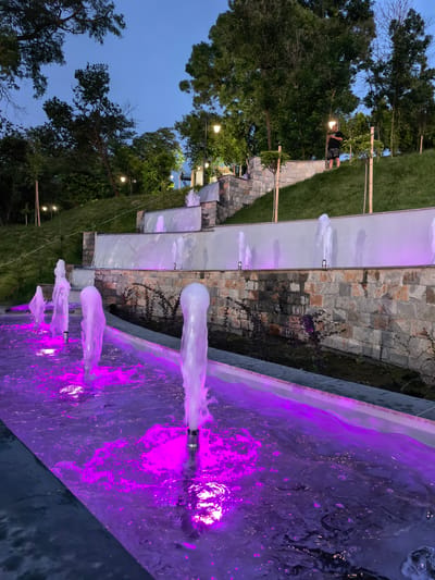 The image shows a tiered fountain with purple lighting in the city of Svishtov, Bulgaria. The fountain has multiple levels with water cascading down. The foreground features several water jets creating plumes of water, illuminated by the purple lights. In the background, a person stands on a grassy slope next to a stone wall, looking at their phone. Trees and streetlights are visible on the upper levels of the park. The sky is a dark twilight blue.