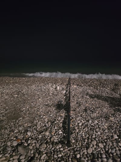 A fishing rod and reel are positioned on a pebble beach at night. The sea is visible in the background with waves breaking. The scene is dark, suggesting it is late at night or early morning. The location is Salerno, Italy.