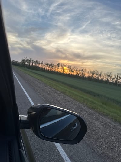 The image depicts a view from inside a moving vehicle, likely a car, driving on a rural road in Fastovetskaya, Russia. The road has a single white dividing line. Adjacent to the road is a strip of green land, bordered by a line of trees silhouetted against the sky at sunset/sunrise. The sky is visible with clouds. The car's side mirror is in the foreground, showing a reflection of the car's interior and the road behind. The asphalt appears to be in good condition. No people are visible in the frame.