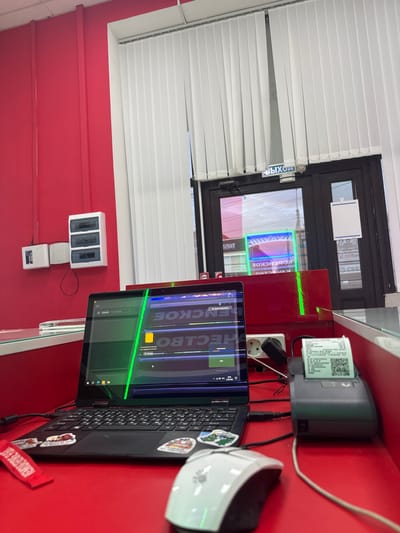In a brightly lit room with red walls, a laptop sits open on a red desk, its screen displaying text in a foreign language and a bright green line. To the right of the laptop is a small receipt printer with a printed receipt, and a white computer mouse is partially visible in the foreground. Behind the desk, a glass partition separates the workspace from a doorway with vertical blinds. A sign above the doorway reads "ВЫХОД" (Exit) in Russian. The room appears to be a commercial space, possibly a shop or office. The presence of electrical outlets and a fuse box on the red wall suggests it is a functional workspace. The reflection in the glass indicates the scene is likely located in Yegorlykskaya, Russia, given the Cyrillic signage.