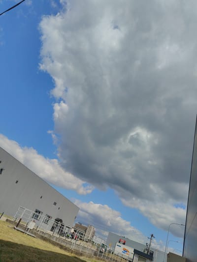 The image depicts an outdoor scene on a cloudy day. The sky is a mix of blue and large, grey clouds. In the foreground, there is a large, white, modern building with a long, slanted roofline. A metal railing runs along the edge of the building. To the left of the building is a patch of dry, yellowish grass. In the middle ground, a road or walkway is visible with some parked bicycles and people walking. Further in the background, more buildings are visible, including one with signage that reads "UNITEL" and "PMK HDI PRZEWIRZTORY STROJOWA". The environment appears to be an industrial or commercial area in Łomna-Las, Poland.