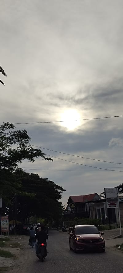The image depicts a street scene in Keudematangkuli, Indonesia, under a cloudy sky with the sun peeking through. A few people are visible on motorcycles, navigating the paved road. A red car is also driving on the road. On the right side of the road, there are buildings with signs, one of which reads "Tukang Gigi BERKAH" and another with a stylized mouth and the word "Berkah". There are also trees and power lines present, indicating an urban or semi-urban environment. The lighting suggests it is either early morning or late afternoon. The overall mood is that of everyday life in a bustling area.