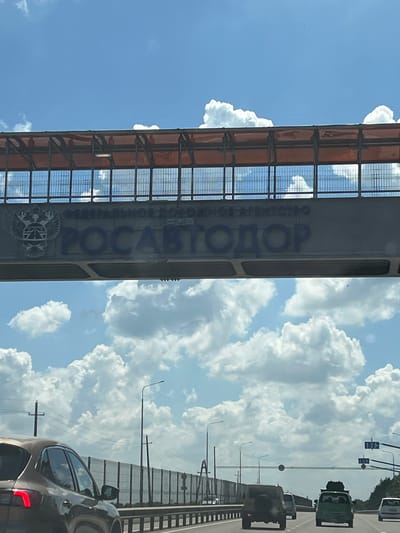 This image, taken in Belozyorny, Russia, depicts a scene from a moving vehicle, likely a car, on a highway on a cloudy day. The perspective is from inside the vehicle, looking upwards and outwards.
The main subject is a large, elevated structure, possibly an overpass or a part of a bridge, running vertically through the frame. This structure is metallic, with a dark gray main beam and a latticed, reddish-orange covering on its upper section, possibly a roof or protective barrier. Large, blue letters are painted on the side of the structure, spelling out "РОСАВТОДОР" (ROSAVTODOR). Below the text, there is an emblem featuring a double-headed eagle with a shield containing a St. Andrew's cross.
The sky is filled with dramatic, fluffy white cumulus clouds against a bright blue sky, suggesting it is daytime, likely mid-afternoon given the intensity of the light. The weather appears warm and potentially humid.
Other objects visible include traffic. Several vehicles are on the highway ahead, including a green truck and a military-style vehicle. The perspective from the camera's car shows the rear of a dark-colored sedan in the foreground, its taillight illuminated. Streetlights and traffic signals are also visible along the roadside.
The scene conveys a sense of travel and movement through a somewhat urban or suburban environment, characterized by infrastructure and vehicular activity under a dynamic sky. There are no people clearly visible as subjects, but the presence of vehicles implies human activity. The overall impression is one of a typical summer day's travel.