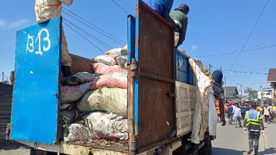 The image depicts a daytime scene on a paved road in Gisenyi, Rwanda, dominated by the rear of a blue and rusted brown flatbed truck. The truck's bed is overloaded with numerous bulging sacks, varying in color (white, pink, green, grey), which appear to contain collected materials or waste. The left rear tailgate of the truck is blue with the white spray-painted marking "B13".

Three individuals are visible in close proximity to the truck:
1.  One person, dressed in a white protective suit with orange trim and green boots, stands on top of the blue tailgate, facing away from the camera.
2.  Another individual, wearing a dark green long-sleeved shirt and dark head covering, is perched on the upper edge of the rusted brown section of the truck's bed, also facing away from the camera. Blue pants are visible.
3.  A third person, wearing a dark blue head covering and a shirt partially covered by a white protective garment, is positioned beside the truck's right rear, holding onto its side.

Further down the street, numerous people are visible, many in civilian clothing, with some wearing reflective vests. One individual in a blue shirt and a yellow-green reflective vest, wearing white gloves, walks away from the truck in the foreground, carrying a long, thin object. Overhead, multiple power lines traverse a clear blue sky. Various buildings are discernible along the street in the background.