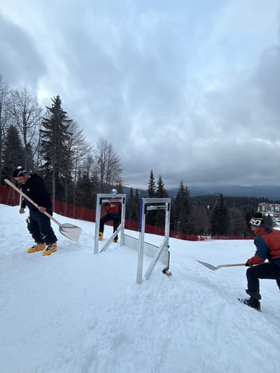 The scene is set on a snow-covered ski slope in Stoikite, Bulgaria, under an overcast sky. Three individuals, all wearing winter ski attire and equipped with skis and boots, are engaged in activities. On the left, a person in a black jacket, black pants, white beanie, and yellow ski boots walks toward the center, carrying a silver-bladed snow shovel with a wooden handle. In the central mid-ground, a person wearing a blue helmet, a dark jacket over an orange top, and yellow ski boots stands between two vertical silver metal frames that are part of a horizontal silver metal structure resting on the snow. This person is looking down at and holding a small electronic device. A ski binding is affixed to the right side of the horizontal structure. To the right, another individual in an orange and blue jacket, dark pants, a dark beanie with a white "RO" logo, and dark ski boots is crouched, actively shoveling snow on the slope with a similar silver-bladed shovel. The background features a dense coniferous forest, with a red safety net delineating the edge of the slope. Distant resort buildings are visible on the far right horizon.