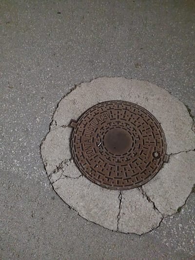 The image shows a manhole cover embedded in a cracked concrete surface, which is surrounded by asphalt. The manhole cover is circular and made of a dark, textured metal, with intricate patterns and some inscribed text that appears to include "ROMA DUCTILACSV" and "EN124 D-400". The concrete around the manhole cover is light gray and shows significant cracks radiating outwards, suggesting wear and tear. The asphalt is a darker gray and has a granular texture. There are no people or animals visible in the image, nor any specific identifiable landmarks. The lighting suggests it might be nighttime or an overcast day, casting subtle shadows. The scene is an urban street or sidewalk environment. This image does not contain enough information to determine its location in Tervel, Bulgaria.