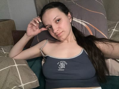 A young woman with dark hair and brown eyes lies on a bed, resting her head on a patterned pillow. She is wearing a dark blue tank top with "ST. GEORGE LONDON" embroidered on the front. Her left arm is bent, and her hand is resting on her forehead. The room appears to be a bedroom, with other pillows and blankets visible in the background. The lighting is dim, suggesting it is nighttime or the room is not well-lit. There are no visible indicators of the specific city of Postoyata, Ukraine, in this image.
