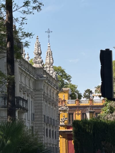 The image displays a sun-drenched architectural scene, likely in Seville, Spain, given the style of the buildings and the bright, clear sky. The perspective is rotated, with the sky appearing on the left and the ground on the right, suggesting the photo was taken from an elevated position or the camera was tilted.
The primary subjects are ornate buildings and their decorative elements. The foreground is dominated by the intricate, weathered stonework of a grand structure, characterized by repetitive patterns and stepped rooflines. Above this, a series of sharp, finial-like spires, also made of stone, punctuate the skyline, one topped with a cross. Behind these, parts of a vibrantly colored building are visible, with yellow walls adorned with red accents and white decorative railings along its upper levels. Lush green trees and foliage are interspersed throughout the scene, with a large, dark green bush prominently featured on the right, and smaller trees and branches framing the lower left. A thick, dark, horizontal log or beam crosses the lower part of the image, partially obscuring the building below.
The bright sunlight indicates it is likely daytime, possibly midday or early afternoon, given the strong shadows cast. The clear blue sky suggests fair weather. There is no visible text in the image. The overall impression is one of historical grandeur and architectural beauty, bathed in the warm light of a sunny day.