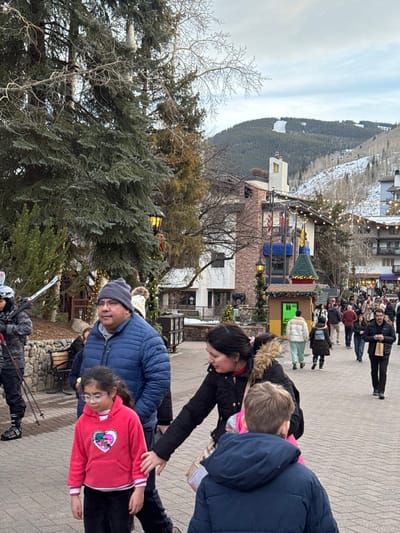 A mountain village scene features pedestrians on a cobblestone path. In the foreground, a group of people is adjusting a child's clothing. Behind them, a man in a blue jacket and a girl in pink stand. A skier with skis is also visible. Buildings decorated with lights and Christmas decorations line the path. A snow-covered mountain is in the background.