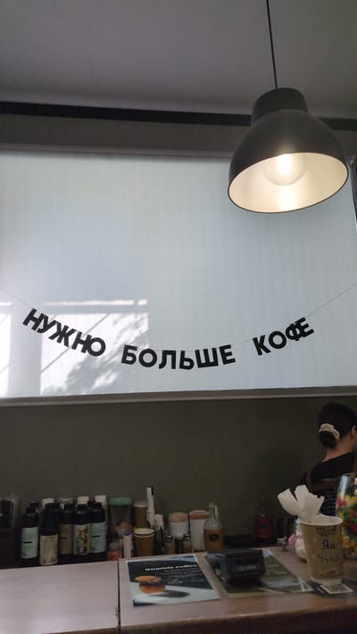 The image captures a cafe interior in Bor, Russia. The primary subject is a wide shot of a counter area and a wall adorned with a banner. On the left side of the frame, a large, dome-shaped pendant lamp with a bright bulb hangs, casting a warm light. To its right, a white roller blind covers a window, with shadows of leaves or blinds visible on it.
A banner with black, block letters is strung across the blind, reading "НУЖНО БОЛЬШЕ КОФЕ" which translates to "MORE COFFEE NEEDED." The letters are arranged in a curve, with "НУЖНО" on the left, arching upwards towards "КОФЕ" on the right.
On the right side of the image, the counter displays various items. A person, mostly out of focus and facing away from the camera, is visible near the top right. In front of them are several objects, including what appear to be small cups, bottles, and possibly jars of candy or condiments. One cup, in the upper right, has handwritten text that is partially legible. Below this, a collection of bottles and cups are neatly arranged on shelves. A small sign with the text "Bumble coffee" is visible on the counter.
The overall impression is a casual, possibly bustling, cafe environment. The lighting suggests it could be daytime, with the shadows on the blind indicating ambient light from outside. The setting appears to be indoors, likely a modern cafe.