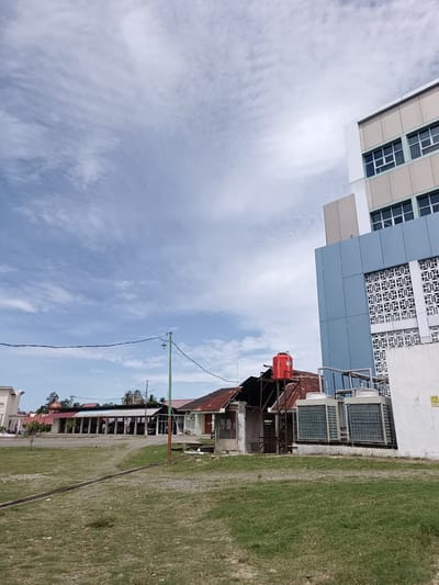 The image depicts an outdoor scene in the city of Mesjid Reuntoh, Indonesia. The sky is partly cloudy with a light blue hue. In the foreground, a grassy area with some gravel and a drainage grate is visible. Several buildings are present, including a modern, multi-story building with blue and beige facades and numerous windows. Adjacent to this structure is an older, single-story building with a rusty corrugated metal roof and what appears to be a red water tank on top. Two large, industrial air conditioning units are situated outside the newer building, near the older structure. In the middle ground, a series of long, low buildings with white pillars and dark roofs are lined up, possibly forming a covered walkway or storage area. A green lamppost stands near the center of the frame, with black wires strung between poles. The overall scene suggests a mix of modern and older architecture within an urban or semi-urban environment. There are no people visible in the image.