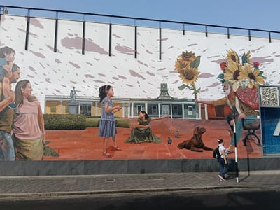 A mural is painted on a wall with a depiction of a town square. A woman stands next to the wall, walking on the sidewalk, and passing a pole. The mural contains images of people, a dog, sunflowers, and a building labeled "Biblioteca." There are also pigeons on the "ground" of the mural. The top of the wall has a horizontal metal railing. This mural is located in the city of Barranco, Peru.
