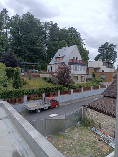 The image shows a residential street scene in Doksy, Czechia, with houses, vegetation, and a red truck. A gray sky with clouds suggests overcast weather. In the foreground, a concrete ledge and a metal fence are visible. A red flatbed truck with a gray cargo bed is parked on the asphalt street. The truck appears to contain some construction materials, possibly including power tools.
Behind the truck, a low gray wall with a red wooden fence runs along the property line. Beyond the fence, a sloped yard is covered with green hedges and trees. A modern-looking house with white walls and a gray roof is situated on a higher level, featuring multiple balconies and large windows. A covered outdoor seating area with furniture is visible on a wooden deck adjacent to this house.
Further down the street, another building with a mix of white and wood-paneled sections and a flat roof is present. The street itself is paved with a textured gray surface and an asphalt section. A sign with the word "POZOR" (Czech for "CAUTION") is visible near the bottom right of the frame, next to a house with a tiled brown roof and stone walls. Vegetation, including trees and bushes, surrounds the houses and properties. The overall impression is of a suburban or semi-rural residential area with evidence of ongoing construction or maintenance.