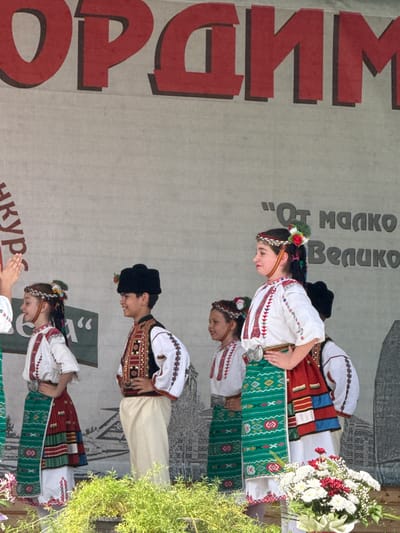 The image depicts a group of children in traditional Bulgarian folk costumes performing on a stage. In the foreground, several children, including girls in embroidered white blouses, colorful skirts, and headbands adorned with flowers, and a boy in a black fur hat, embroidered vest, and white pants, are the main subjects. They appear to be engaged in a dance or presentation, with some individuals looking to the side and others facing forward. The backdrop features a large banner with red lettering. Part of the banner reads "Орхим" in large red capital letters, and to the right, smaller text in black is visible, including "От Малко" and "Велико". The setting suggests an outdoor event, possibly a festival or cultural celebration, held in the city of Pordim, Bulgaria, given the prominent display of the city's name on the banner. Lush green foliage is visible at the bottom of the image, hinting at a natural or park-like environment.