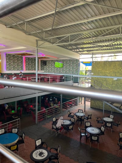 The image captures a bright, daytime scene inside a multi-level, open-air establishment, likely a restaurant or bar. Natural light streams in through the extensive corrugated metal roof structure supported by industrial beams, indicating clear weather.
On the upper level, walls are decorated with a striking green leaf-patterned wallpaper or fabric, augmented by vibrant pink/purple and yellow strip lighting. A television screen, displaying green imagery, is mounted on one of these patterned walls. A blue sign prominently displays the word "WASHROOMS." Several individuals are seated at tables with red chairs on this level, engaged in leisure activities.
A sturdy metallic railing forms a barrier, overlooking the lower ground floor seating area. This lower level is characterized by a distinctive floor paved with hexagonal tiles in alternating red and dark gray/black. Numerous black chairs surround an array of round and square tables. Many of the round tables feature the "TUSKER" brand logo, complete with its elephant graphic, a strong indicator of the location in Kenya. A few people are visible seated at these tables, suggesting a casual dining or drinking environment.
The overall atmosphere is relaxed, designed for socializing and entertainment, with the presence of TVs and multiple seating options. Visible text includes "WASHROOMS" and the "TUSKER" brand logo. The architecture and branding align with the provided context of Kimorori, Kenya.