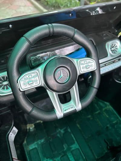 This media file is a close-up, top-down view of the interior of a black toy Mercedes-Benz G-Wagon, specifically focusing on the steering wheel and dashboard area. The steering wheel is black with silver accents and features the Mercedes-Benz logo in the center. There are buttons and controls on the steering wheel, some with text labels. One of the silver accents on the steering wheel has "AMG" written on it. The dashboard has a digital display that resembles a speedometer and tachometer. To the right of the steering wheel, a dark green textured plastic interior panel is visible. The setting appears to be outdoors, as evidenced by the blurred green foliage and a hint of pavement or a walkway in the upper left corner, suggesting a yard or driveway in Pîrlița, Moldova. The lighting suggests it is daytime, likely with overcast skies or in the shade, as there are no strong shadows. The overall impression is one of a detailed, realistic toy car interior. There is a white label with a barcode and some text visible on the green plastic panel on the right.