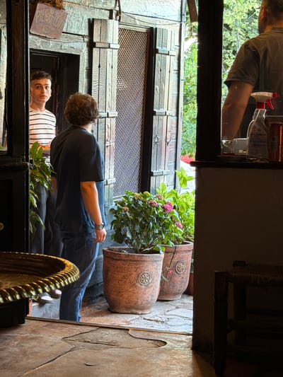 Two young men are seen through an open doorway in what appears to be an outdoor cafe or restaurant setting. The man on the left, facing the camera, wears a striped t-shirt and has short, dark hair. He is holding a piece of food. The man on the right, with curly brown hair, is seen from the back, wearing a dark t-shirt and blue jeans. He is looking into the distance. In the foreground, two terracotta pots containing flowering plants are visible. The ground is paved with stone. The doorway itself is made of weathered wood and has decorative shutters. In the background, greenery suggests a garden or courtyard. The sign above the doorway reads "PİERLOT." This establishment is located in the Eyüpsultan district of Istanbul, Turkey.