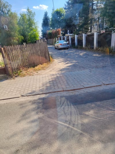The image depicts a street scene in Jaworowa, Poland, under a bright, sunny sky with scattered clouds, suggesting daytime. A silver sedan is parked on a gravel surface adjacent to a paved walkway. The walkway is constructed of interlocking concrete pavers, laid in a stretcher bond pattern, bordered by a curb. Beyond the walkway, there's a dirt path leading to a wooden fence made of vertical planks.
In the background, a concrete and metal fence with white square posts separates properties. Trees with green foliage line the properties behind the fence, and a building is partially visible. A red and white sign with the word "SOLID" is affixed to the fence. The overall impression is a quiet residential or semi-rural area bathed in sunlight, with shadows cast across the paved surfaces, indicating the time of day. There are no people visible in the scene.