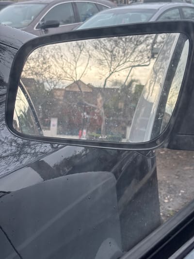 A car's side mirror reflects a building and bare tree branches. The mirror frame is black, and the reflective surface is speckled with water droplets. A portion of the car's hood, also black and wet, is visible on the right side of the frame. Another car is reflected adjacent to the side mirror.