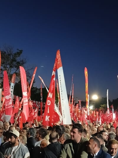The image shows a large crowd of people gathered in an open-air space in Silivri, Türkiye, at the coordinates 41.071, 28.244. The crowd is holding red flags with white lettering, including the words "NDIK" and "CHP". Some of the flags are also decorated with a white star and crescent moon. The crowd is densely packed, and it is dark outside. There are a few streetlights illuminating the scene. Many of the individuals in the crowd are wearing casual clothing. The people are looking towards the front of the crowd, some with smiles and excitement. They are participating in an event, potentially a political rally.