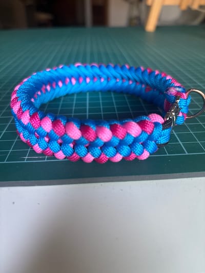 A close-up shot shows a blue and pink braided bracelet with a silver clasp and ring. The bracelet is lying on a green cutting mat with a grid pattern. The background is blurred and shows a wooden surface and indistinct objects. There is no indication of the city of Coslada, Spain, or any people present in the image.