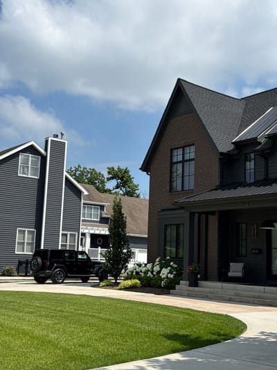 The image depicts a suburban street scene in Naperville, Illinois, United States of America. In the foreground, a meticulously manicured bright green lawn curves away from the viewer. A light-colored concrete driveway intersects the lawn, leading to two residences and a parked black Jeep Wrangler.

The house on the left is a two-story structure with dark gray siding and white trim around multiple windows. A prominent, tall chimney with white siding and a metal cap rises from the left side of this house.

The house on the right is a larger, more contemporary residence constructed with dark brick and dark siding. It features a dark-colored asphalt shingle roof with some modern metal roofing elements. A prominent dark gray porch with multiple steps leads up to the entrance. The house number "804" is visible on the facade above the entrance. A small black planter with a pink flower and a single chair are visible on the porch.

Behind the Jeep, white-flowered bushes and a dark green evergreen tree are planted. In the background, more trees and the roofs of other houses are visible. The sky is a clear blue with scattered white clouds. The overall impression is one of a well-maintained and affluent neighborhood on a sunny day.